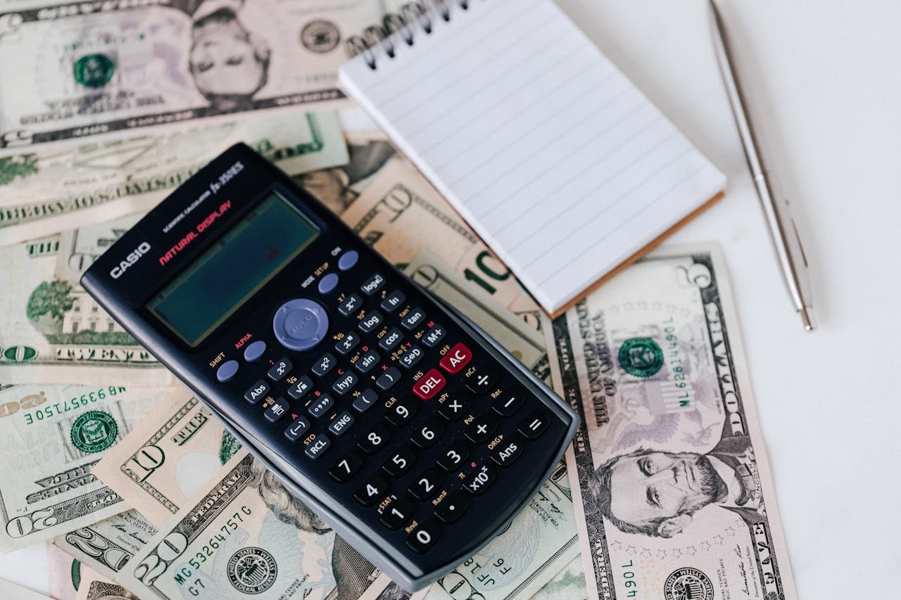 From above electronic calculator and notepad placed over United States dollar bills together with metallic pen for budget planning and calculation